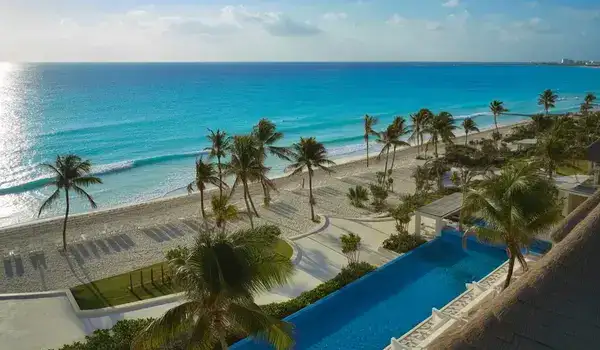 Aerial view of an exclusive Cancun resort with white sandy beaches, turquoise ocean, beachfront villas, palm trees, and infinity pools.